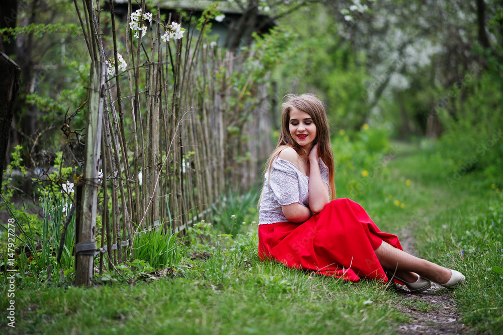 Portrait of sitting beautiful girl with red lips at spring blossom garden on green grass, wear on red dress and white blouse. Portrait of sitting beautiful girl with red lips at spring blossom garden on green grass, wear on red dress and white blouse.