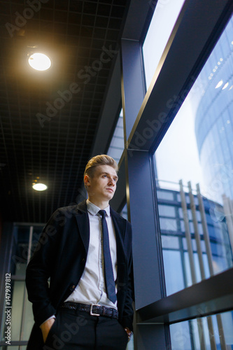 Low angle view of pensive young entrepreneur with hands in pockets standing by panoramic window of spacious office corridor, portrait shot