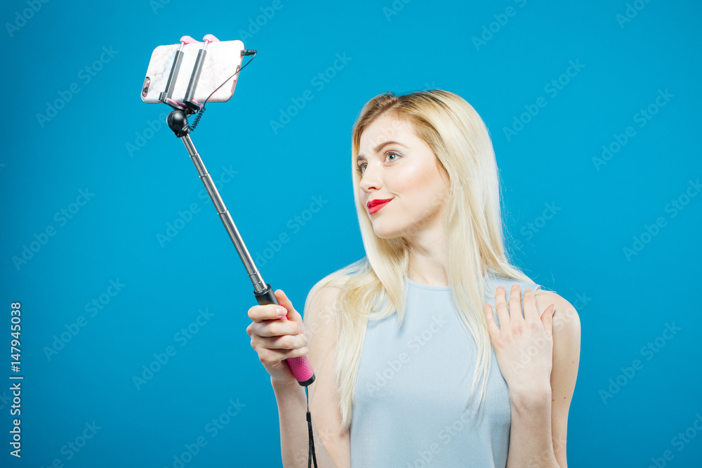 Portrait of Cute Blonde with Sensual Lips Photographing Herself. Smiling Girl Using Selfie Stick to Take a Photo on Blue Background. Portrait of Cute Blonde with Sensual Lips Photographing Herself. Smiling Girl Using Selfie Stick to Take a Photo on Blue Background.