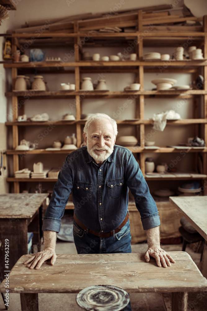 Front view of senior potter standing and leaning on table against shelves with pottery goods at workshop Front view of senior potter standing and leaning on table against shelves with pottery goods at workshop