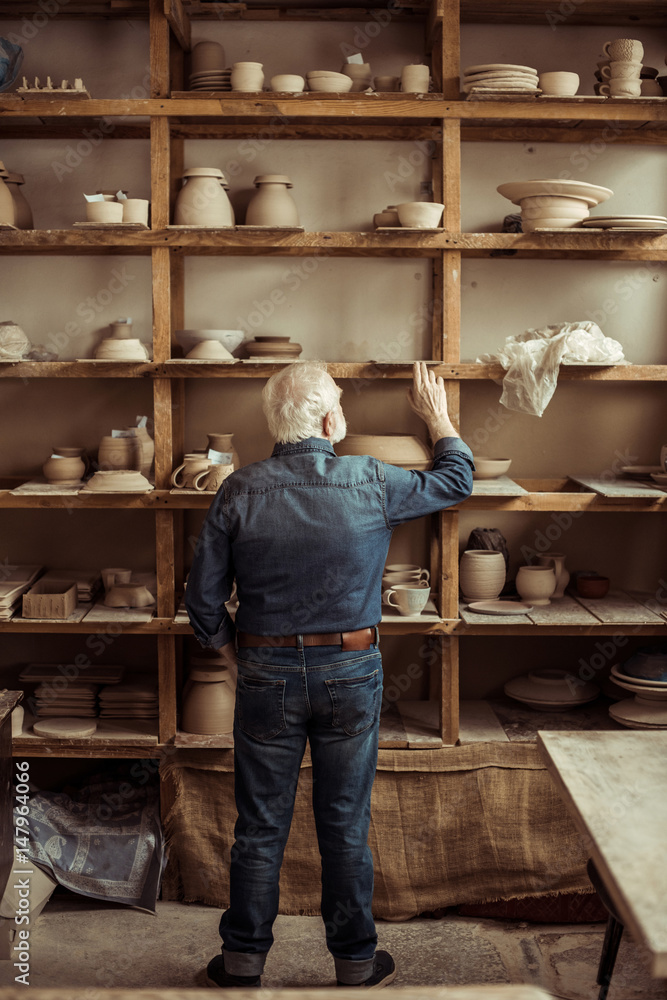 Rear view of senior potter standing near shelves with pottery goods and searching something at workshop