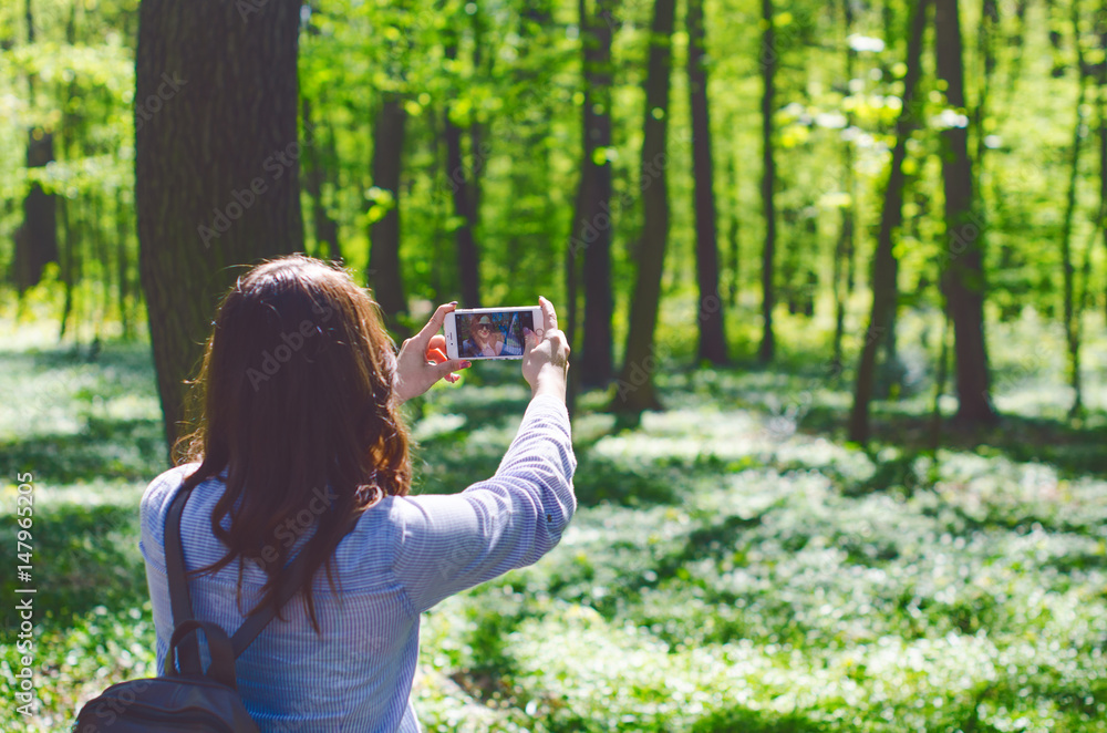 Young woman take a photo with smart phone on a pathway in the forest. Beautiful woman with bag. Nature. Deciduous trees. Spring time. Pleasure weather and sun light