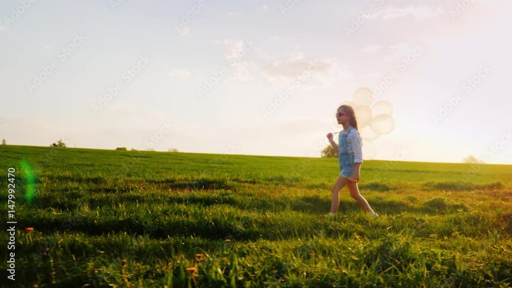 Little girl in pink glasses with balloons. Walking along a country road on a background of a green meadow and a blue sky. Concept - a happy childhood, a childhood dream Little girl in pink glasses with balloons. Walking along a country road on a background of a green meadow and a blue sky. Concept - a happy childhood, a childhood dream