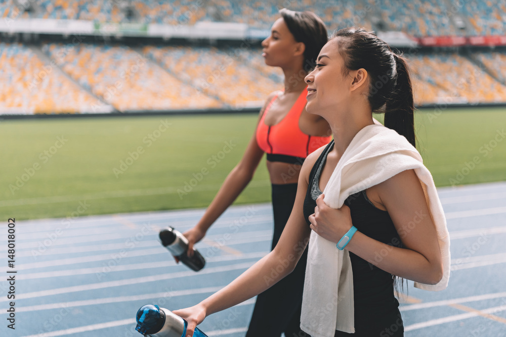 Tired young sportswomen with bottles of water walking on running track stadium