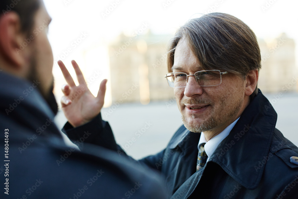 Bearded middle-aged manager looking at his colleague with squinty eyes while talking to him outdoors, over shoulder view