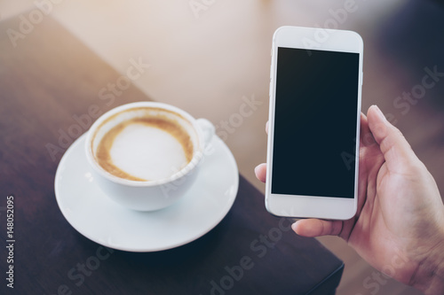 Mockup image of hands holding white mobile phone with blank white screen and hot coffee cup on wooden table in cafe