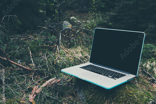 Freelancer laptop computer lost in the forest glade, amid the greenery, summertime. Photo depicts notebook computer in the woods, blurred forest green plants behind. Freelance concept.