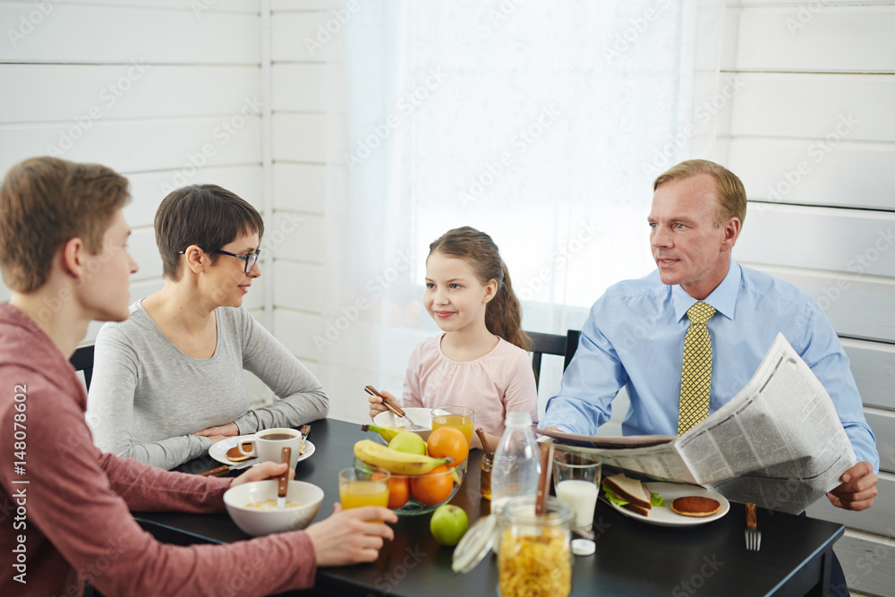 Peaceful breakfast in bosom of family: red-haired father with newspaper in hands talking to his teenage son, little daughter and her short-haired mother enjoying delicious food