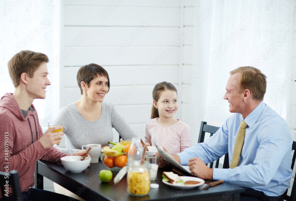 Lovely family moments at breakfast time: pretty middle-aged mother, little daughter and teenage son looking at head of family with wide smiles while he entertaining them with funny story Lovely family moments at breakfast time: pretty middle-aged mother, little daughter and teenage son looking at head of family with wide smiles while he entertaining them with funny story