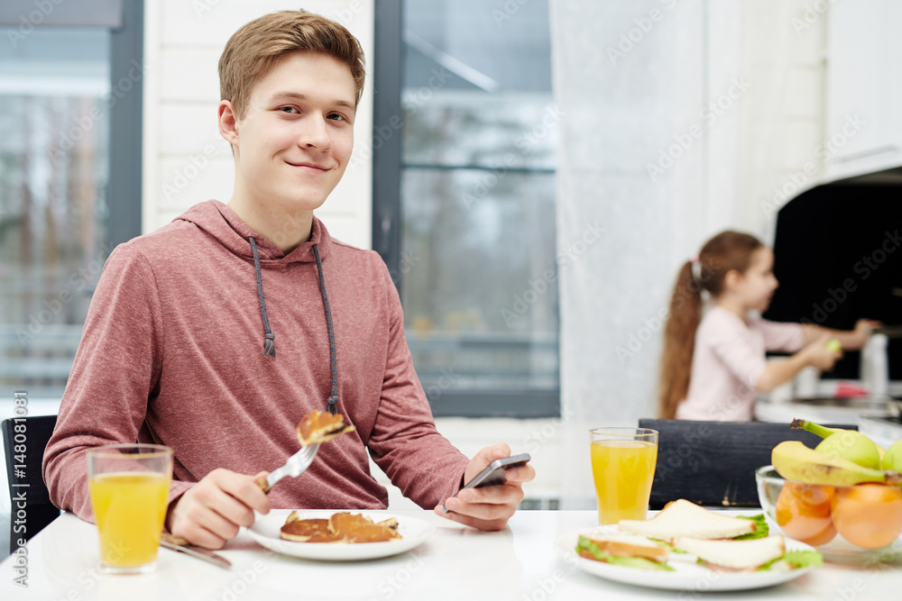 Waist-up portrait of handsome teenage boy looking at camera with wide smile while eating tasty pancakes at breakfast, his little sister standing at sink and washing dishes