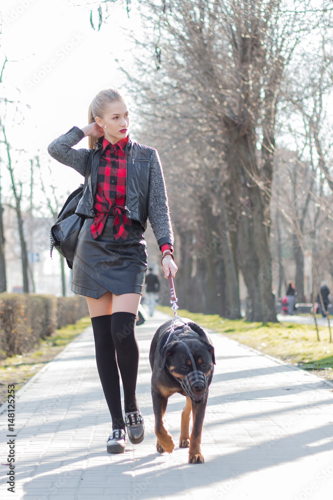 Beautiful young girl walking his dog in a muzzle on a short leash. Dressed in a leather jacket, leather skirt, stockings, and a shirt in a cage.