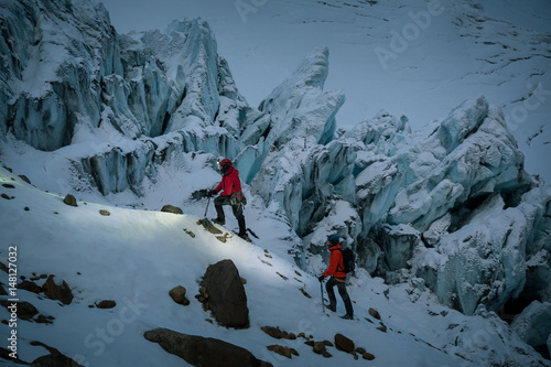 Two people ascending snow covered hillside by rock peaks, Ecuador, South America photo