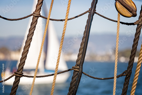 Nautical background. Close up of sailboat ropes, cables and wood pulley in grid pattern. Blurred white sailboat and blue water in the background.