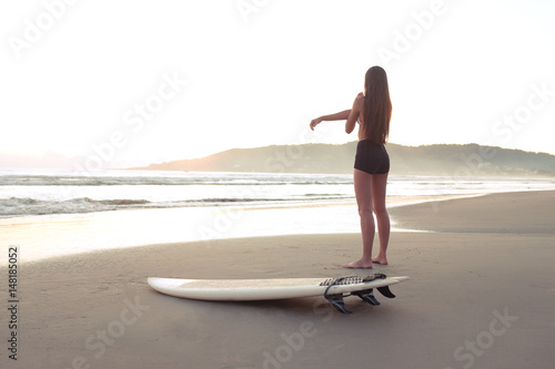 Female surfer warming up and stretching at the beach before entering ocean at sunset or sunrise