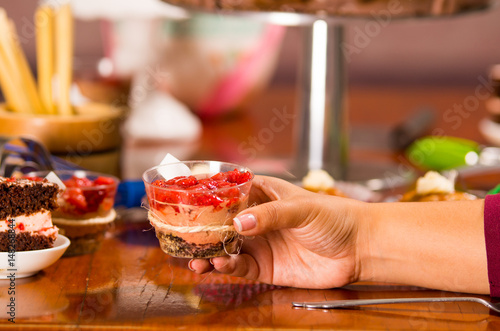 Hand grabbing onto delicious mousse covered in strawberry topping, presented inside small glass, cake pieces and muffins background, pastry concept