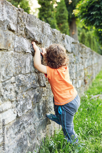 A little boy tries to climb a stone wall at summer evening in city park.