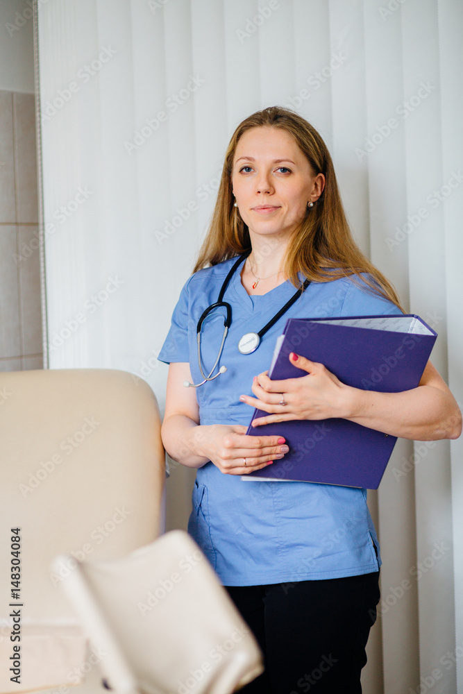 Young woman gynecologist standing behind an armchair and holding blue folder with document Young woman gynecologist standing behind an armchair and holding blue folder with document