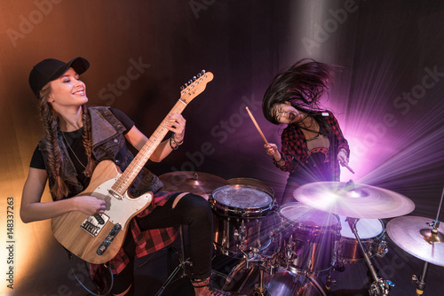 Excited young women with drums set and electric guitar performing rock concert on stage