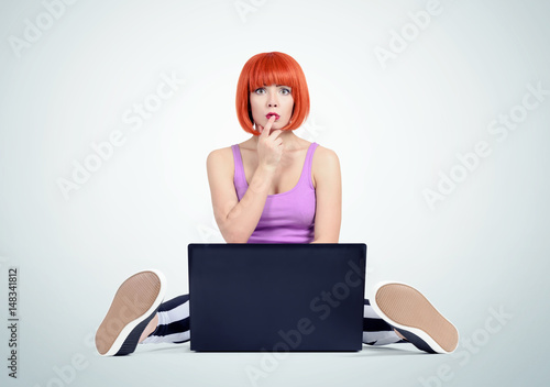 Young red-haired girl sits in front of a laptop pressing her finger to her lips. Oops! 