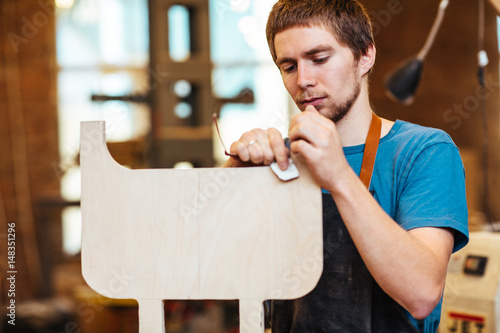 Portrait of focused young man making furniture in carpenters workshop, sanding wooden detail with file photo