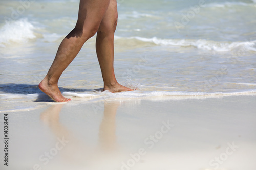 Caucasian tanned woman is walking on the beach. Water and sand on the feet. White sand beach with waves.