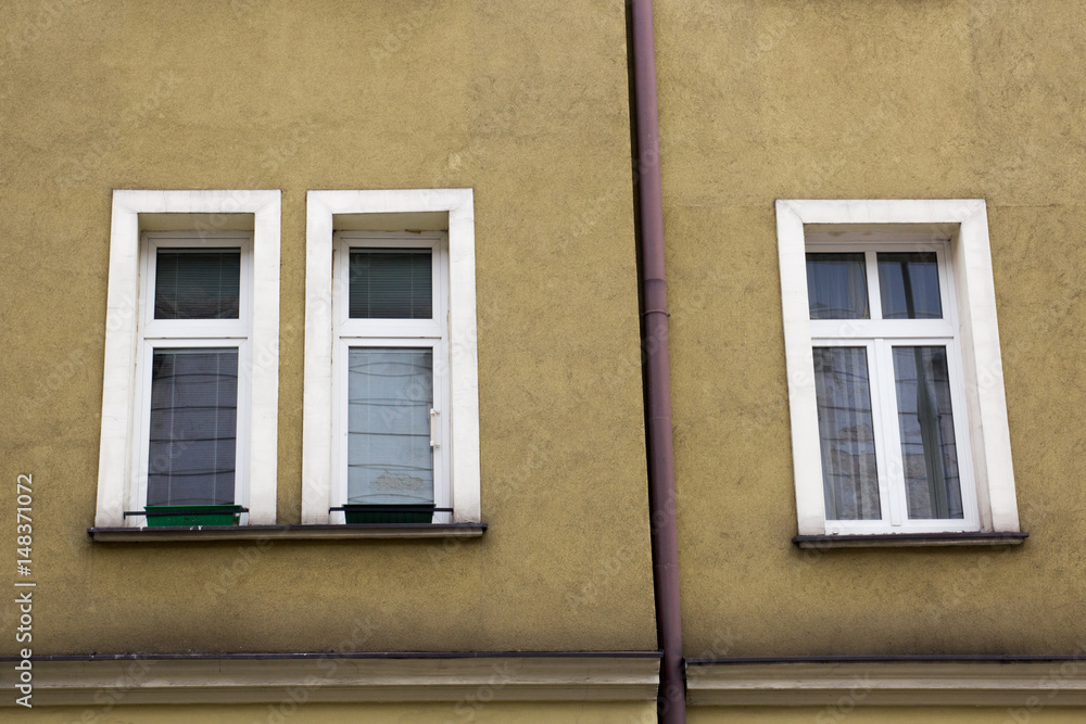 Three white windows on the yellow facade of the house with a drain pipe