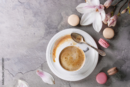 White cup of black coffee, served on white saucer with macaroons biscuits, spoon and magnolia flower blossom branch over gray texture background. Flat lay, space