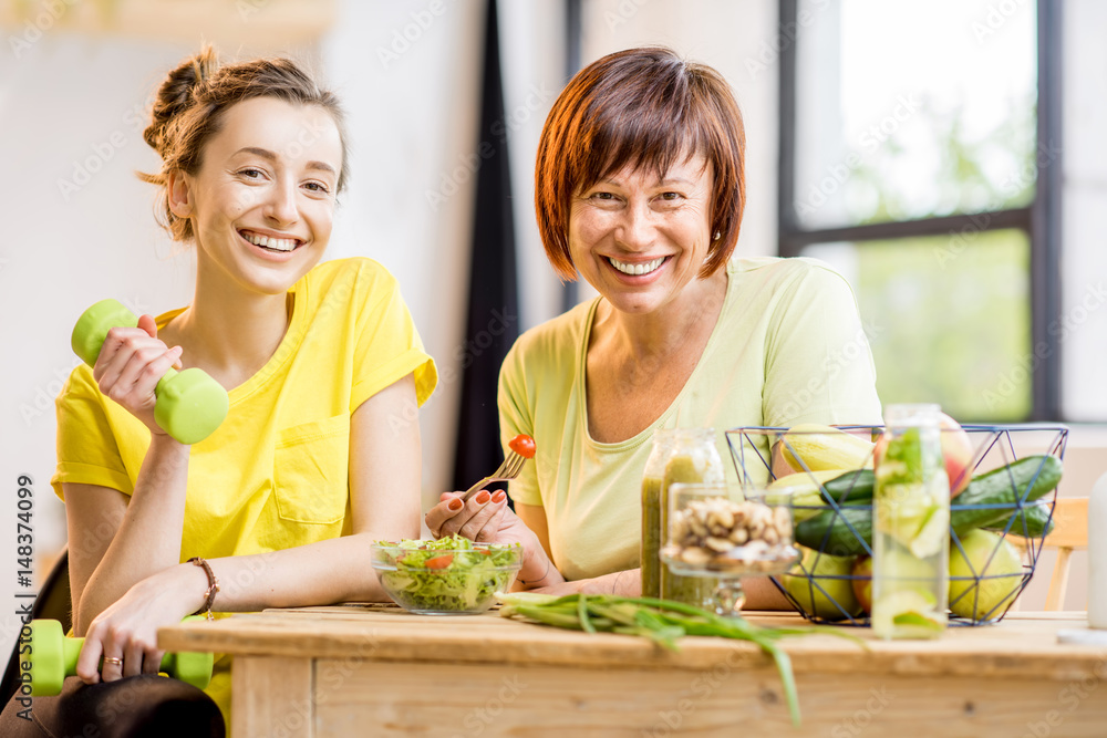 Young and older women sitting with healthy food and fresh drinks after the sports training indoors on the window background