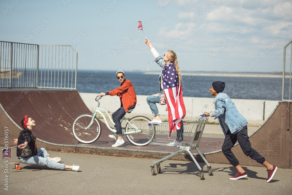 Obraz premium Teenagers group with skateboard, bicycle and shopping cart having fun together and waving american flags at skateboard park