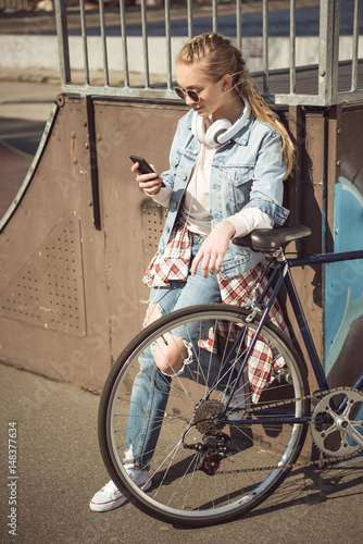 Fototapeta Naklejka Na Ścianę i Meble -  Stylish young blonde girl in sunglasses standing with bmx bike and using smartphone at skatepark