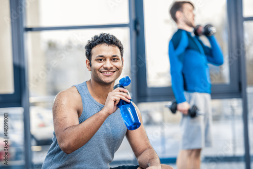 Handsome young man with bottle of water resting at gym, other man workout on blurred background