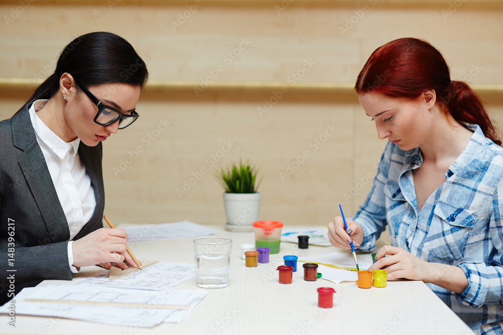 Waist-up portrait of two pretty women concentrated on work: ambitious architect drawing project with pencil and ruler while creative designer sitting opposite her and creating colorful picture