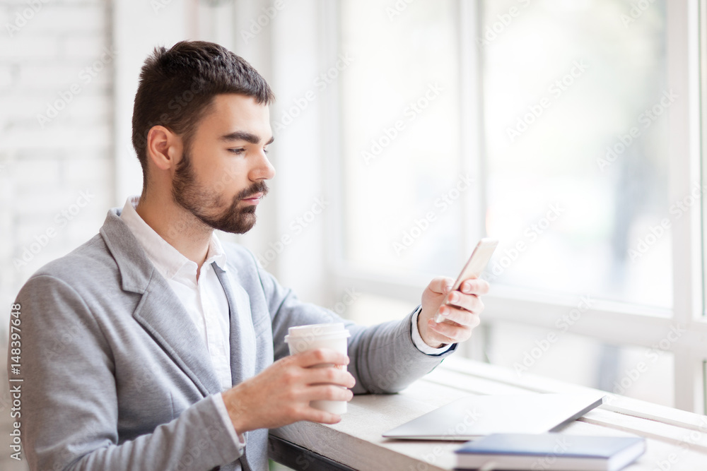 Calm businessman having coffee break in cozy cafe: he sitting at panoramic window with paper cup in hand and texting with his friend, waist-up portrait Calm businessman having coffee break in cozy cafe: he sitting at panoramic window with paper cup in hand and texting with his friend, waist-up portrait