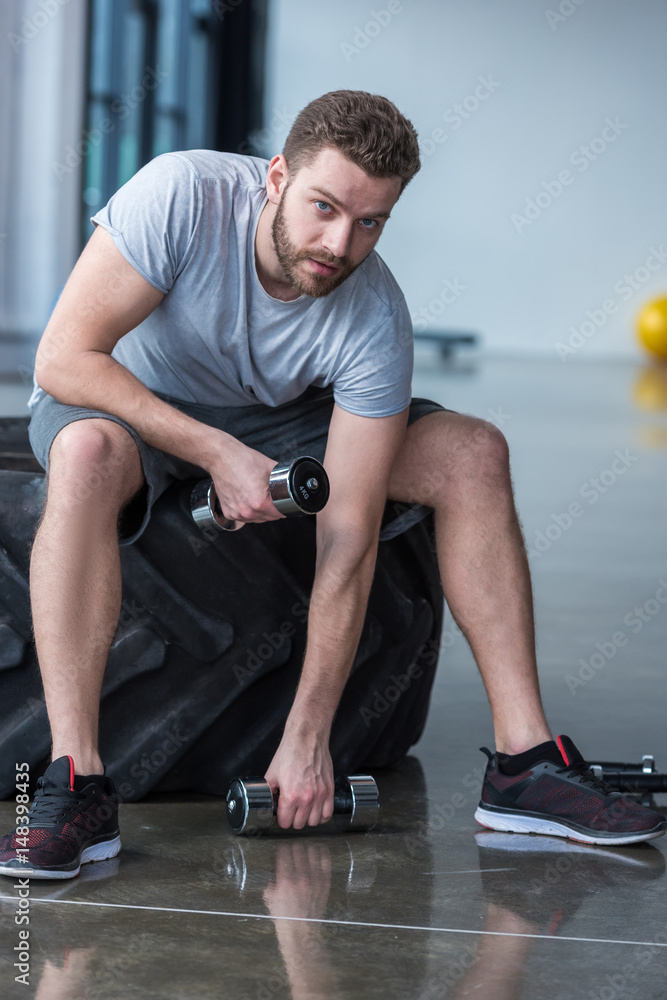 Obraz premium Young man sitting on tyre and exercising with small dumbbells