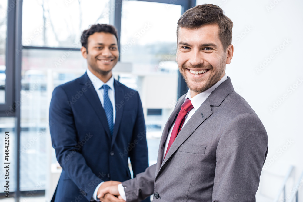 Handsome young businessmen shaking hands and smiling at camera, business concept Handsome young businessmen shaking hands and smiling at camera, business concept