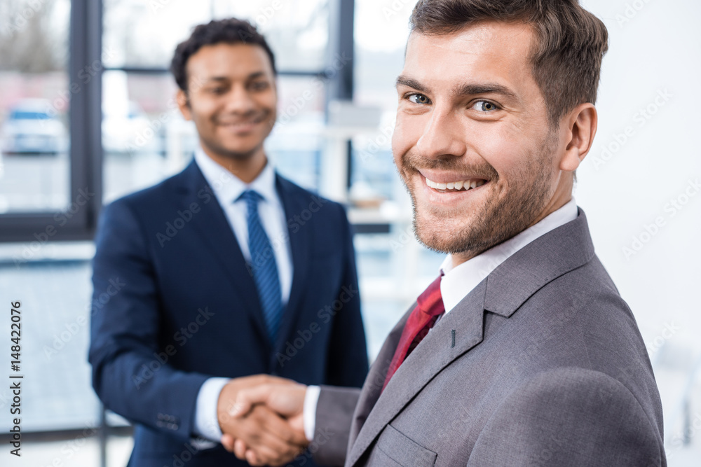 Handsome young businessmen shaking hands and smiling at camera, business concept Handsome young businessmen shaking hands and smiling at camera, business concept