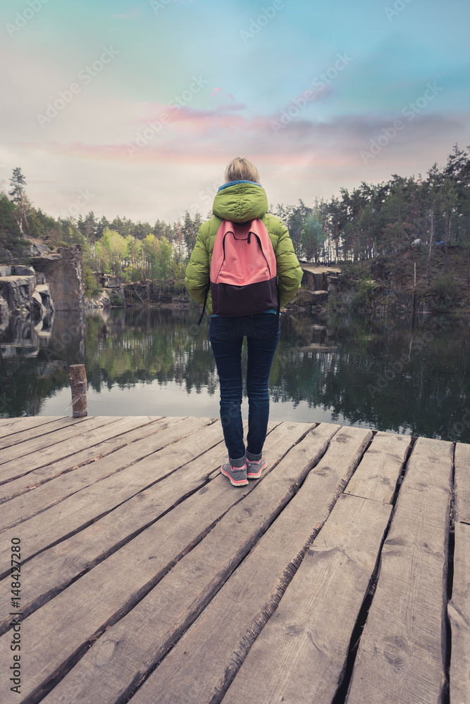 A female traveler in a light green jacket and a pink-brown backpack is standing on a wooden pier near a beautiful lake surrounded by stone rocks in the middle of a pine forest.