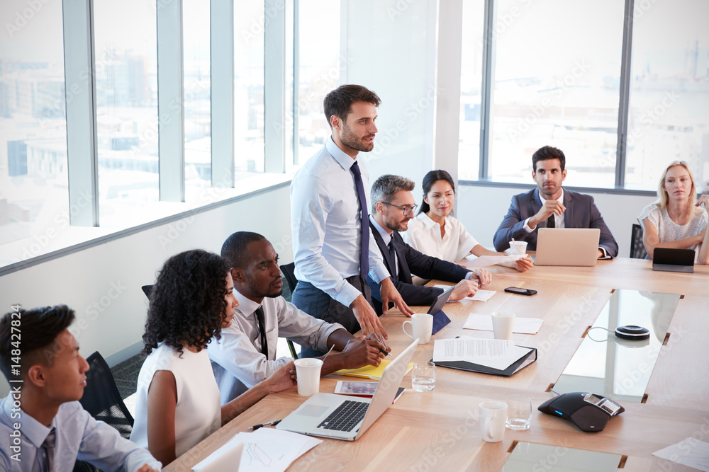 Businessman Stands To Address Meeting Around Board Table Businessman Stands To Address Meeting Around Board Table