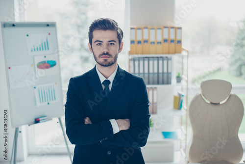 Concept of successful young confident businessman. Concentrated man with crossed arms standing in front of shelves full of folders and board with graphs