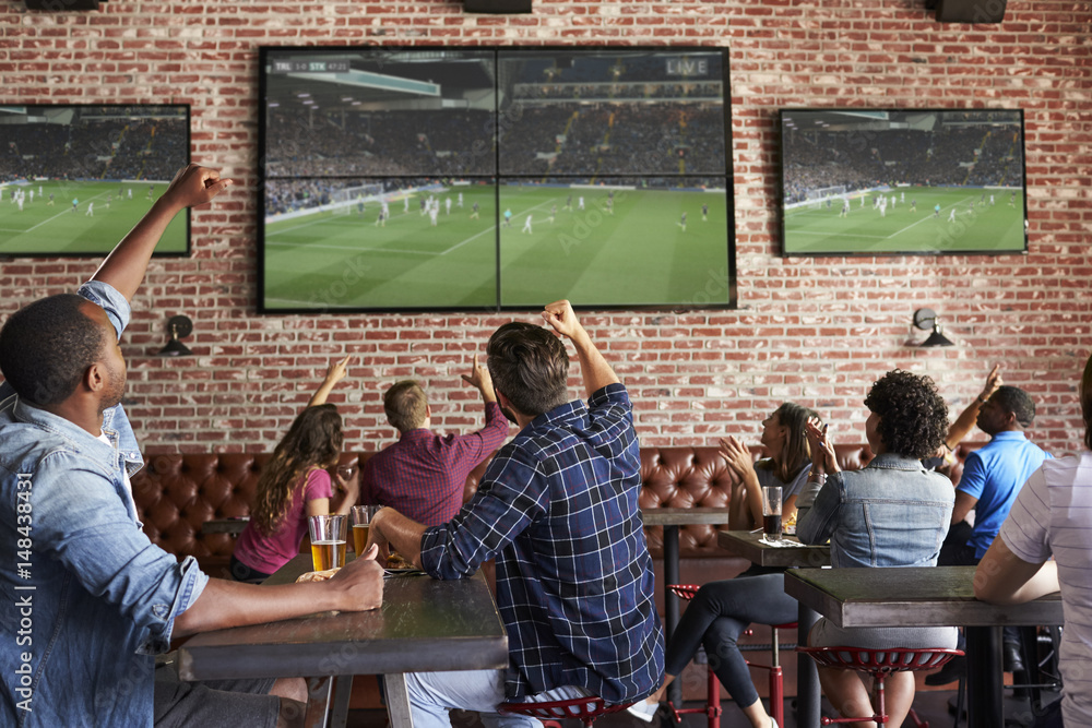 Friends Watching Game In Sports Bar On Screens Celebrating - obrazy, fototapety, plakaty 