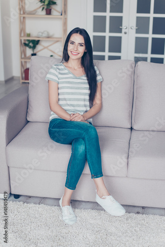 Free Download Vertical portrait of cute pretty smiling woman sitting with crossed legs on a sofa at home - GFXLoad