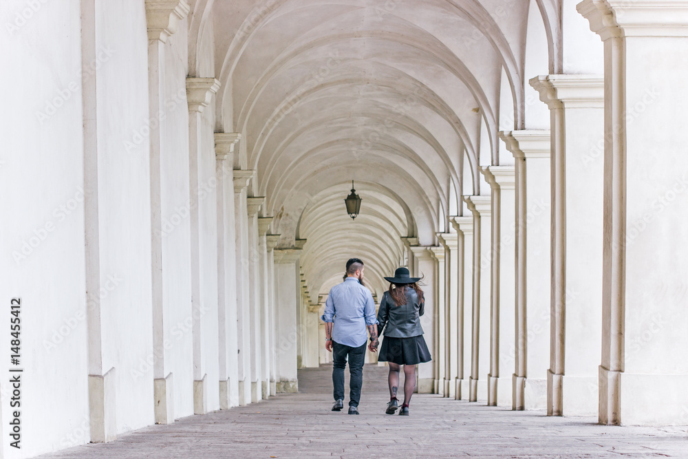 Happy young couple holding hands and walking together under the arches. Young modern hipster couple in love.