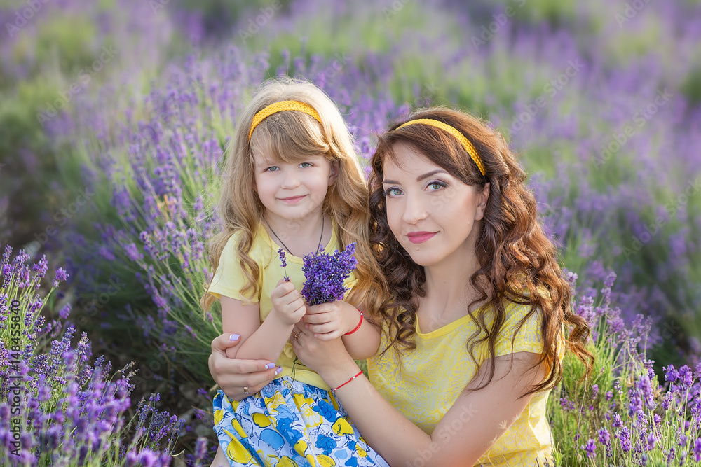 Young mother with young daughter smiling on the field of lavender .Daughter sitting on mother hands.Girl in colorful dress and mother in dark blue dress.