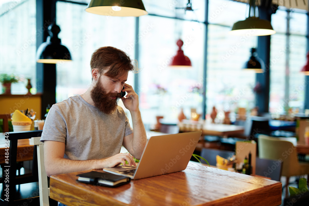 Obraz premium Busy bearded manager sitting in cyber cafe and talking to his business partner on mobile phone, modern laptop and notebook located on wooden table, waist-up portrait