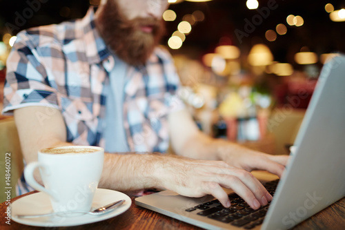 Close-up shot of male hands typing on laptop keyboard, cup of delicious cappuccino standing on wooden table
