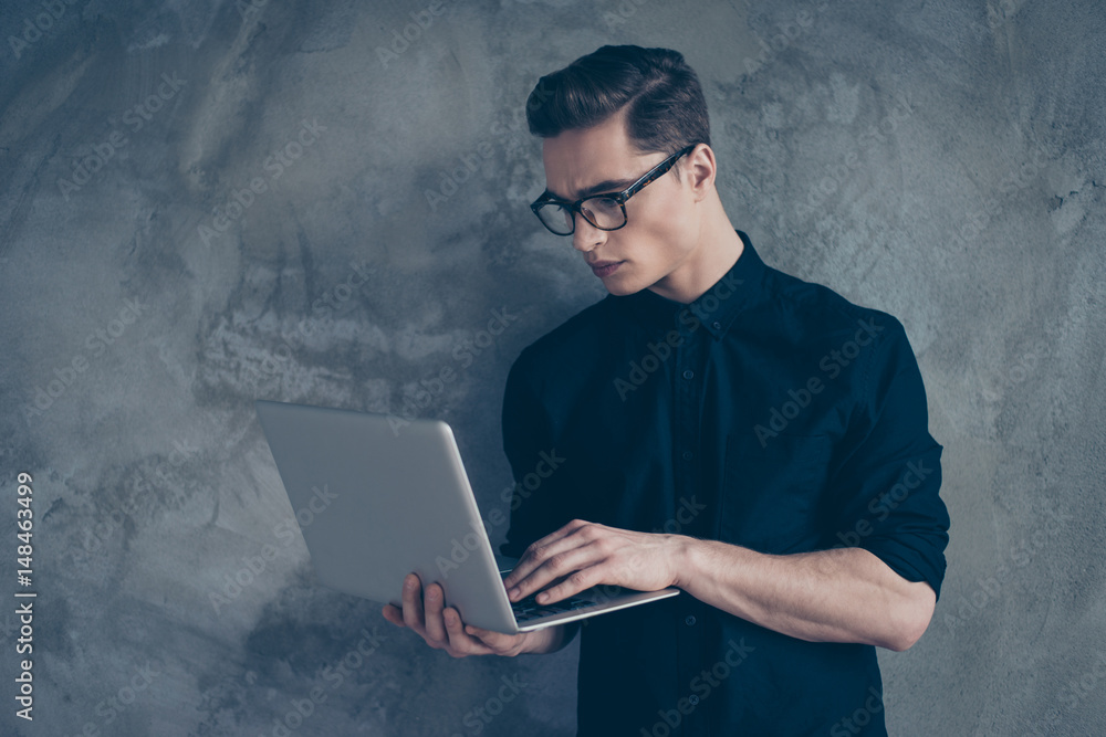 Side view of young attractive businessman in black shirt and glasses, using his laptop on the grey background Side view of young attractive businessman in black shirt and glasses, using his laptop on the grey background