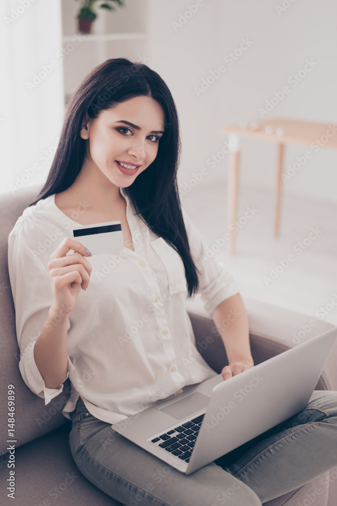 Concept of online shopping and payment. Vertical portrait of young smiling woman sitting on a sofa at home, using a computer and showing credit card. Concept of online shopping and payment. Vertical portrait of young smiling woman sitting on a sofa at home, using a computer and showing credit card.