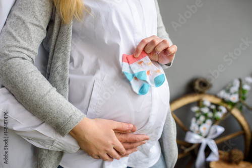 Pregnant woman playing with baby booties on her pregnant belly on last month of pregnancy. Husband touching his wife belly, hands