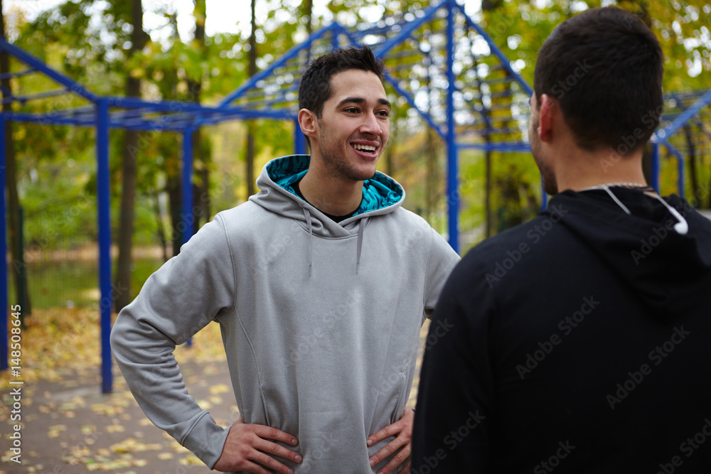 Smiling young sportsman with hands on hips talking to his friend while taking short break during training, waist-up portrait Smiling young sportsman with hands on hips talking to his friend while taking short break during training, waist-up portrait