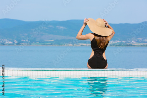 Back view of Brunette woman in swimsuit and hat sitting near the pool